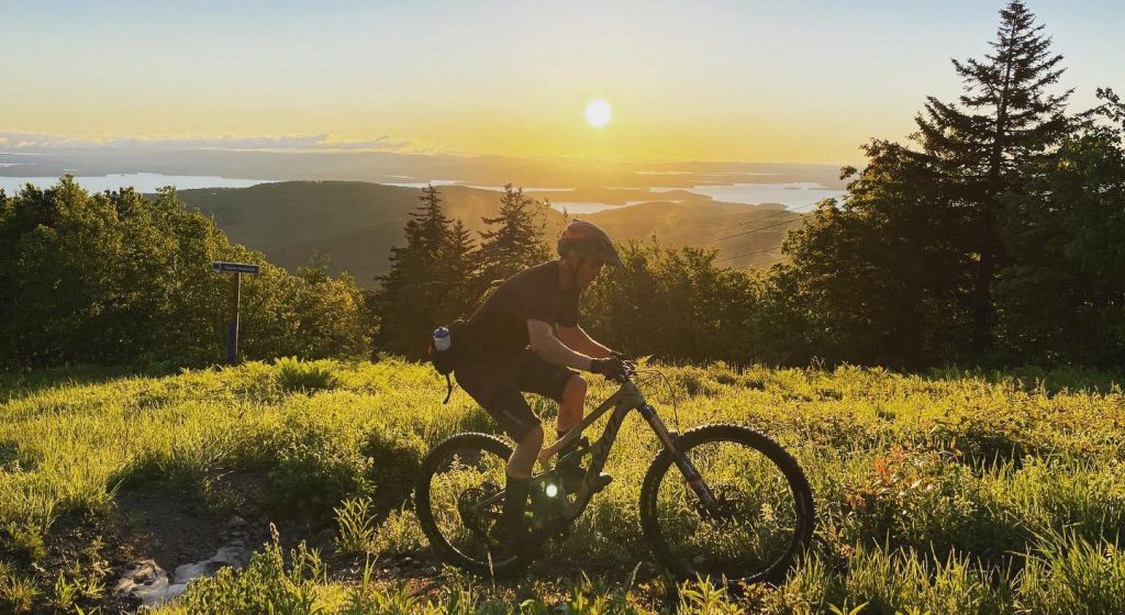 Man riding a mountain bike through a field with lake and sunrise in the background